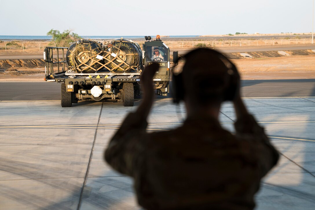 An airman, shown from behind, gestures with his arms to the operator of a loading device on a flightline.