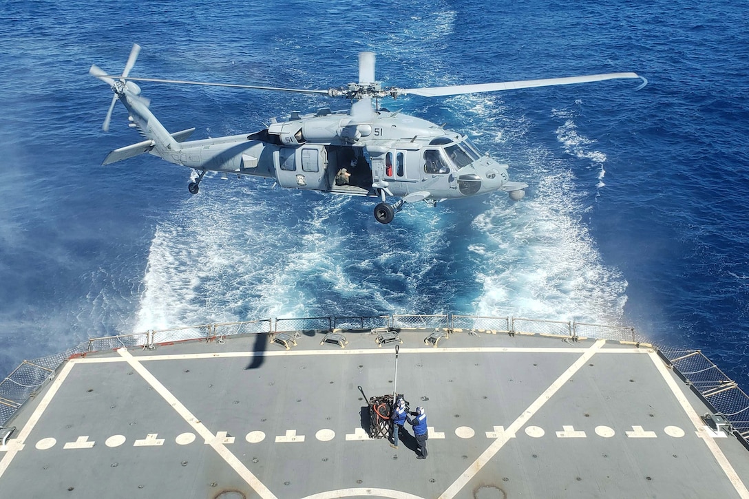 A helicopter hovers above sailors on the deck of a ship.