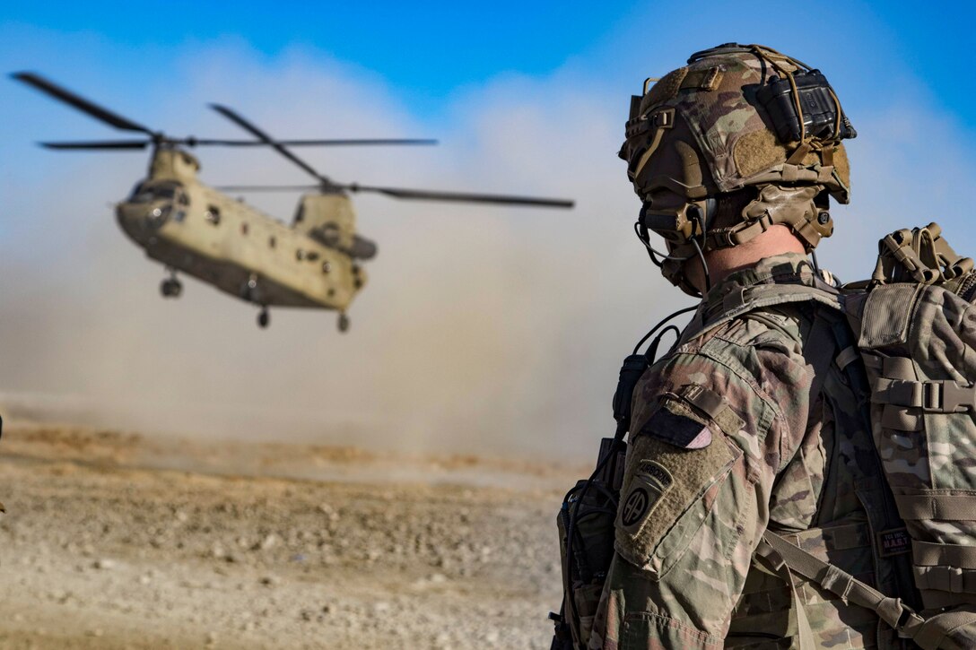 A soldier watches as a helicopter approaches the ground, kicking up dirt.