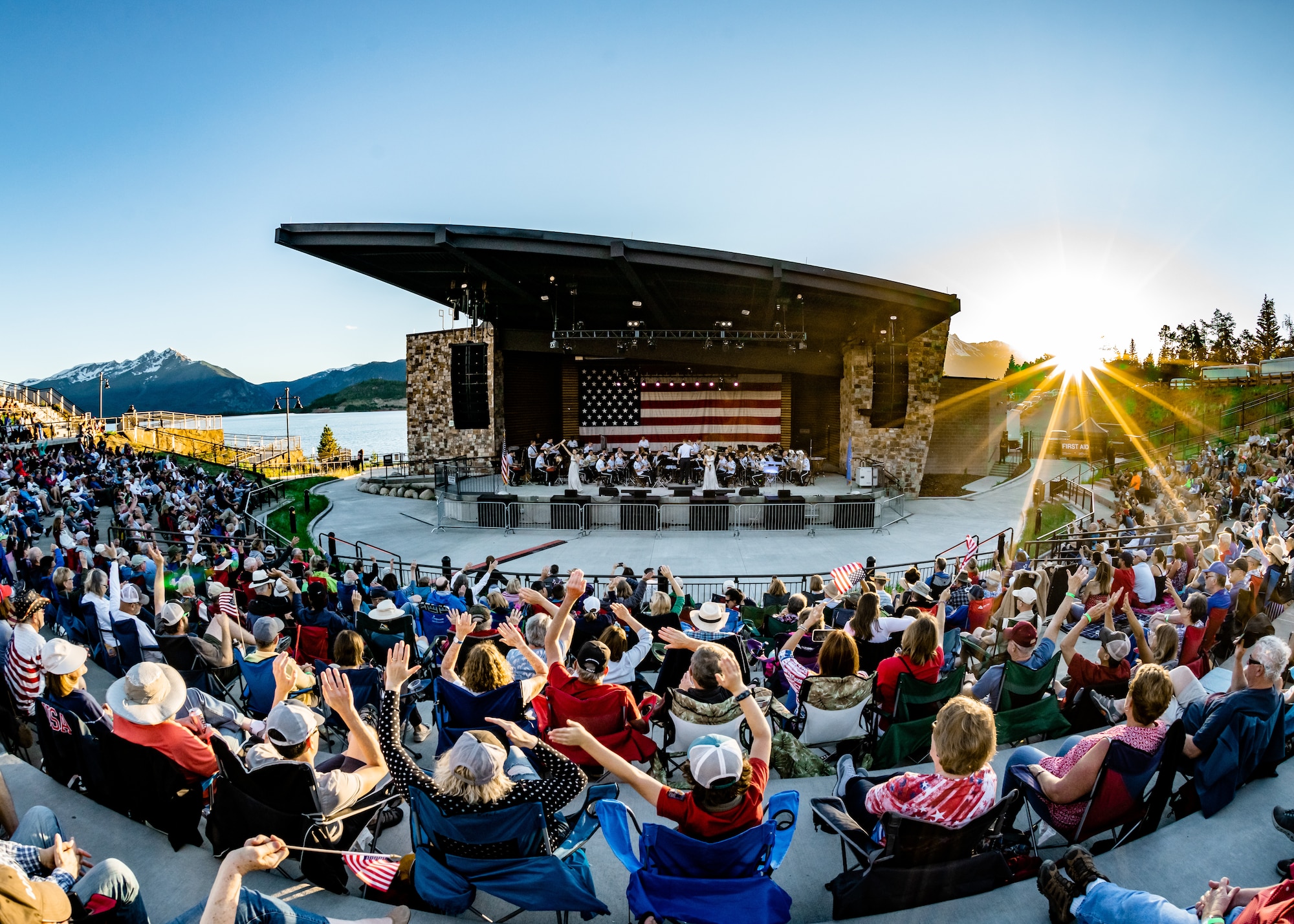 A crowd of 6,000 celebrated the Fourth of July with the Air Force Academy Band at the Dillon Amphitheater in Dillon, CO in 2019.  The crowd waves their arms and small flags in the air as the band plays on a large stage with an American flag backdrop, surrounded by blue snowy mountains, a broad reservoir, and the golden setting sun. Photo by Jenise Jensen.