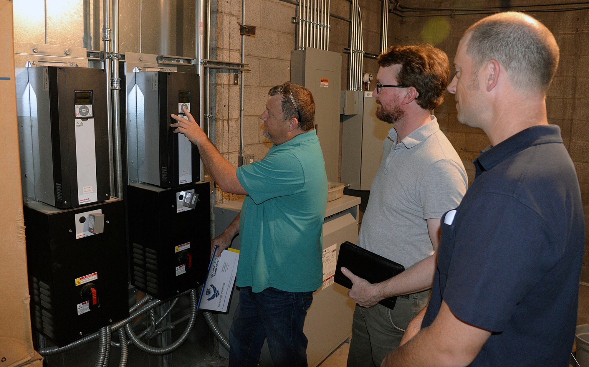 Shane Jepson, facility manager for building 220, shows ISO 50001 auditors Jonathan Clark and Rob Ellis, newly installed variable frequency drive monitors located on the wall of a utility room.