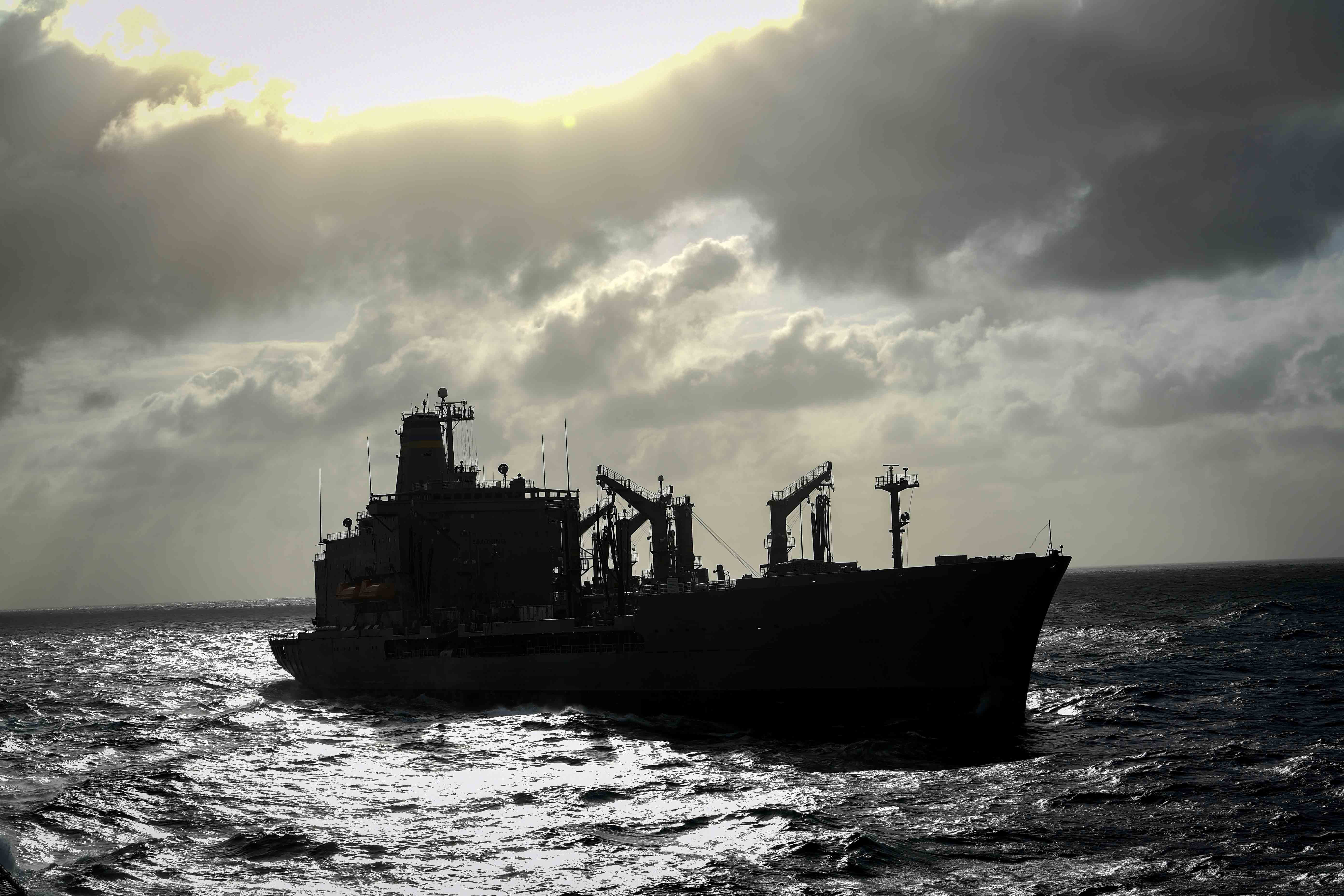 A military ship  on the water is silhouetted against the sky.