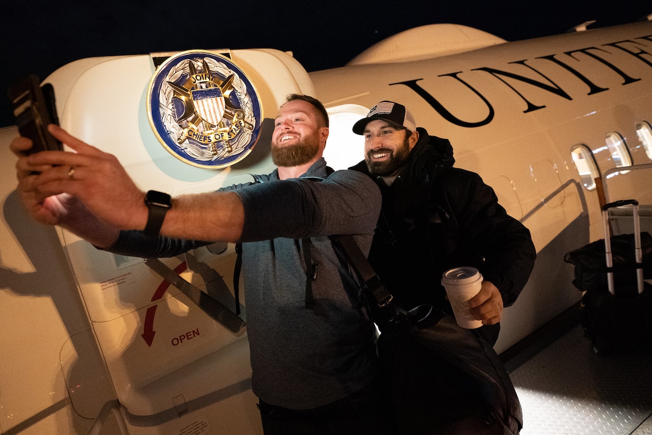 Two bearded men take a selfie at the door or a military aircraft that bears the seal of the Joint Chiefs of Staff on its door.