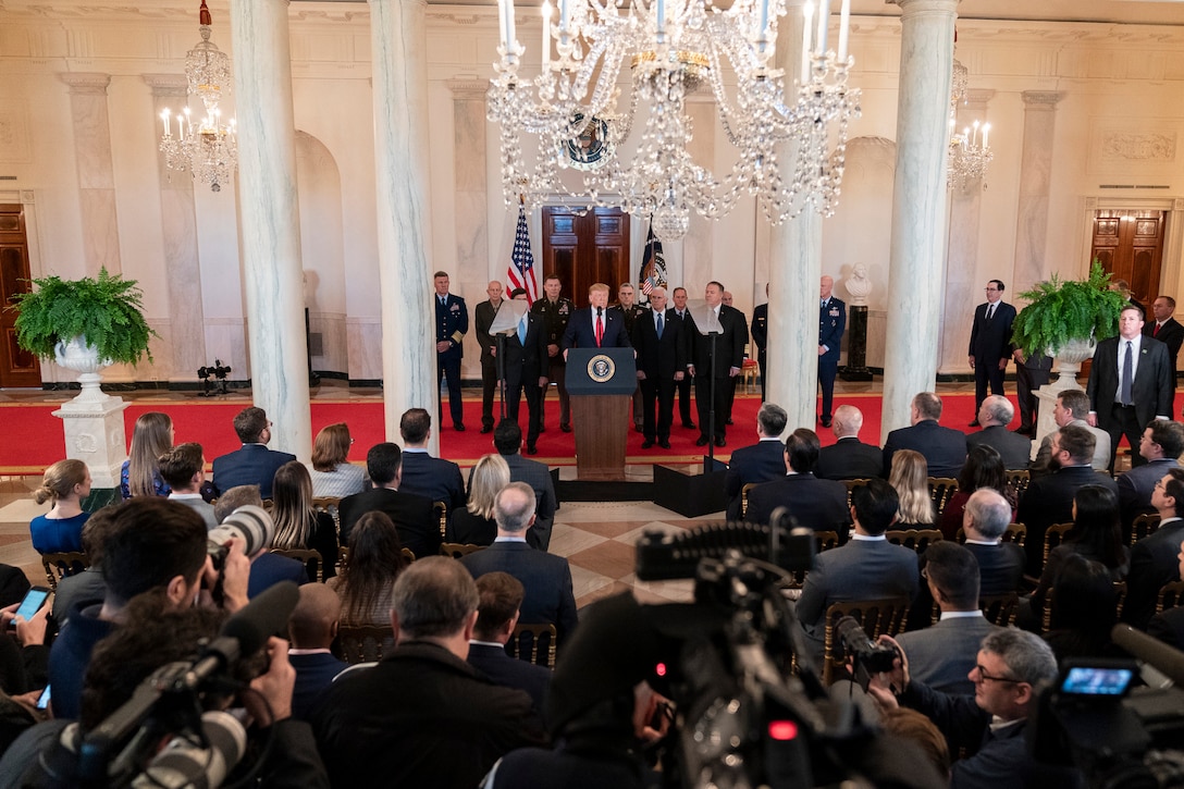 President Donald J. Trump stands at a podium speaking to the press, with senior advisors standing behind him.