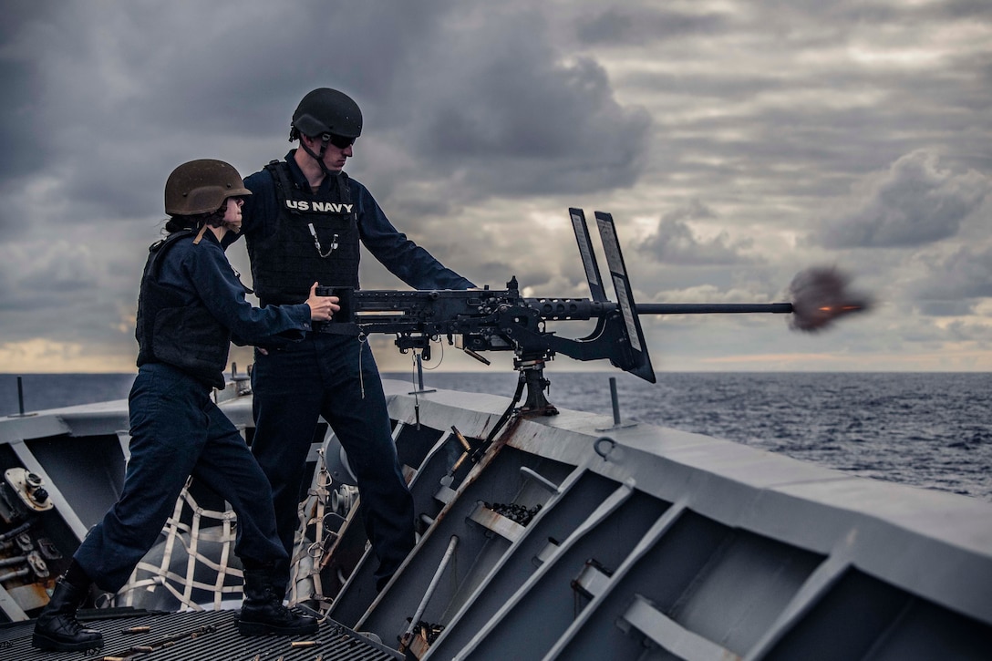 Two sailors stand next to each other as one fires a machine gun.