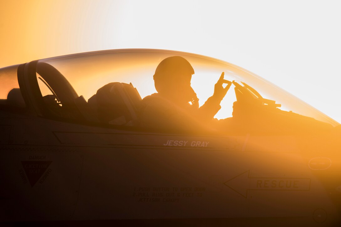 A pilot in a cockpit, shown in silhouette and illuminated by orange light, gives a hand signal.