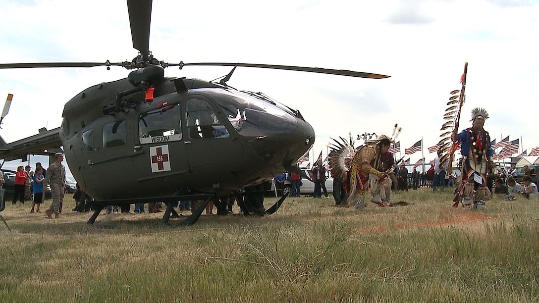Two men dressed in Native American garb dance near a UH-72 Lakota helicopter in a field.