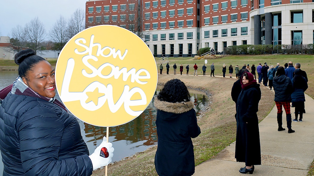 Woman in blue coat holds up a "Show Some Love" sign during an event at the McNamara Headquarters Complex pond at the agency’s Fort Belvoir, Virginia, headquarters.