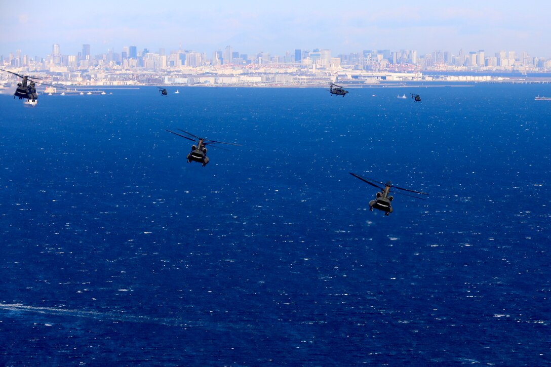 A group of military helicopters fly above the water with a coastline behind them.