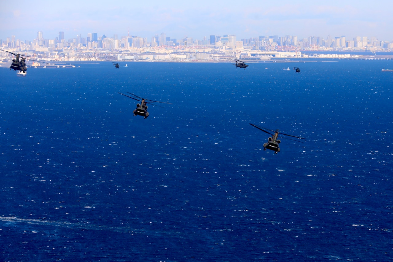 A group of military helicopters fly above the water with a coastline behind them.