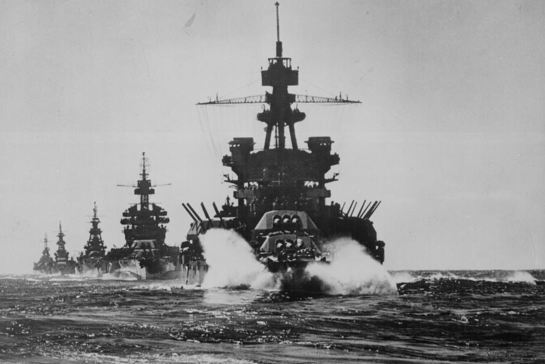 A line of large military ships with guns pointed in the air moves through the ocean.