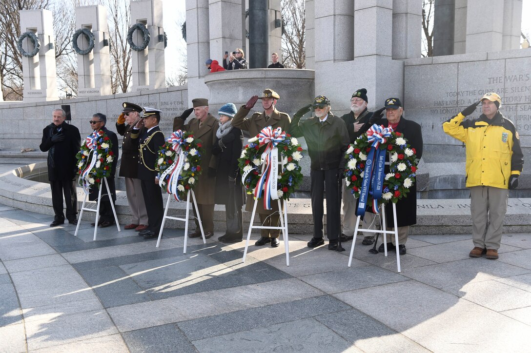 Eleven people stand near a military memorial and behind four floral wreaths. Many are saluting.