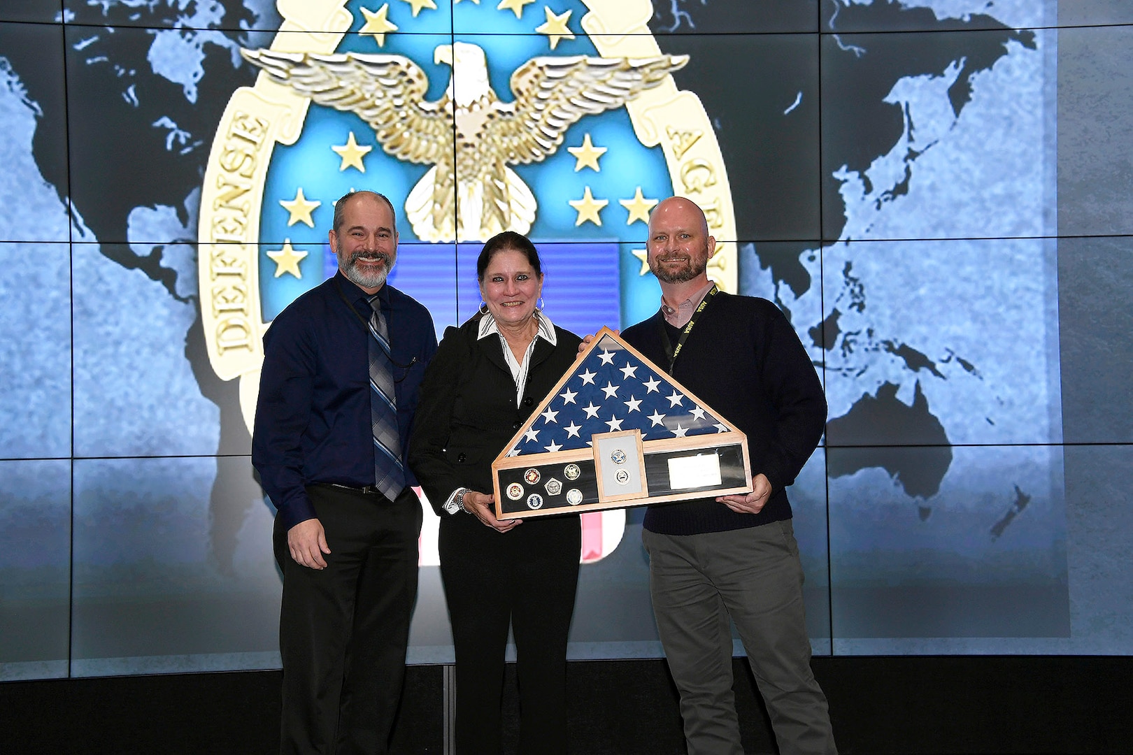 Two men and a woman stand on stage with a flag in a box