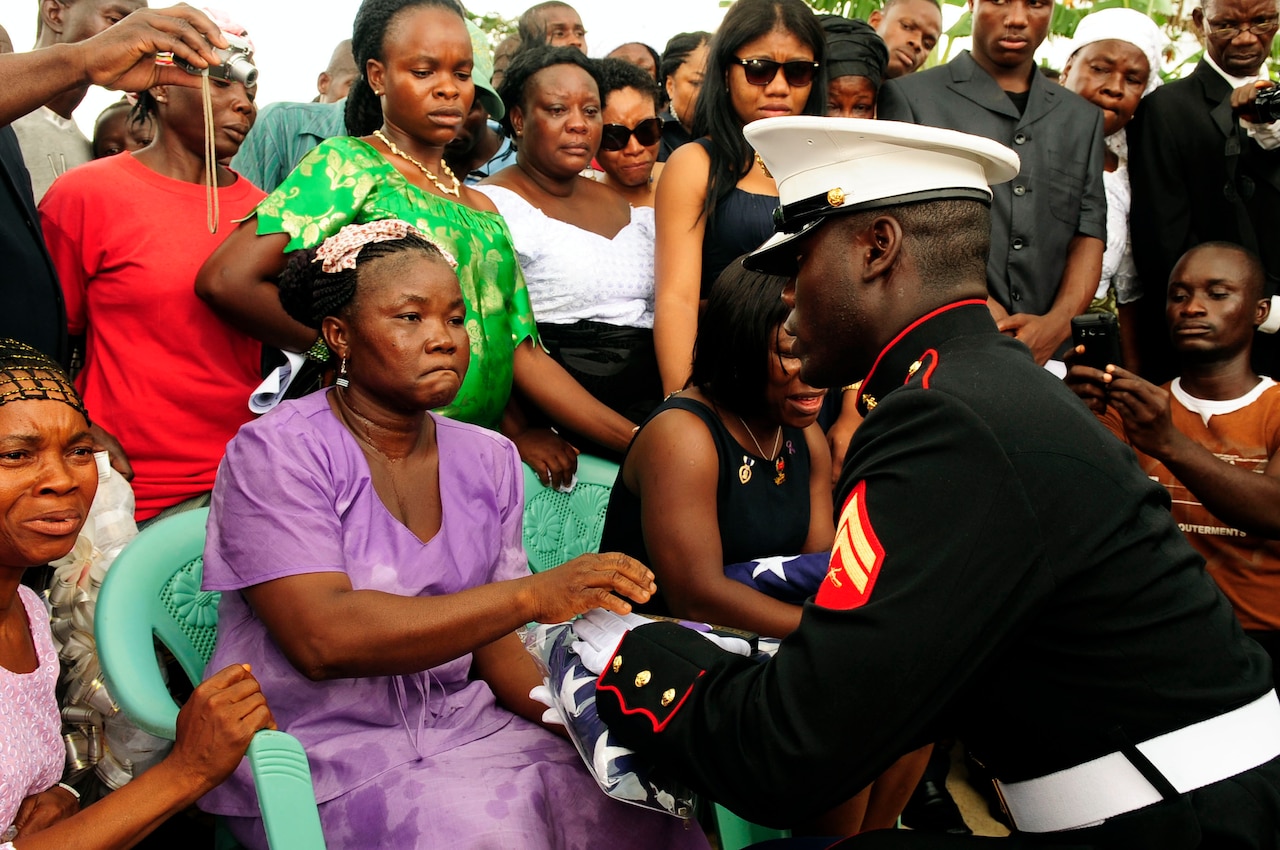 A U.S. Marine hands a folded American flag to a grieving woman sitting in a chair; she is surrounded by other people.