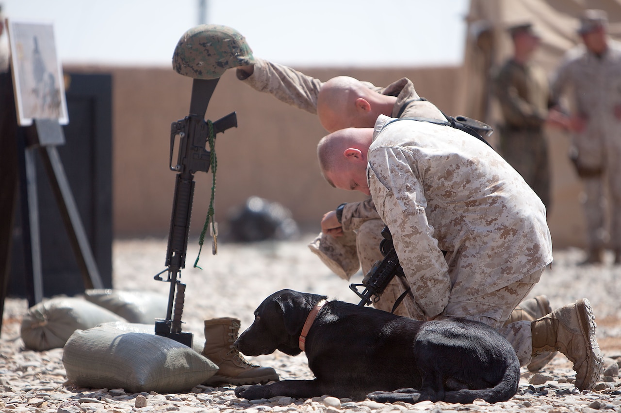 Two Marines kneel and bow their heads in front of an empty boot, helmet and rifle that are propped up in remembrance of a fallen Marine. Beside them, a black dog sits on the ground.