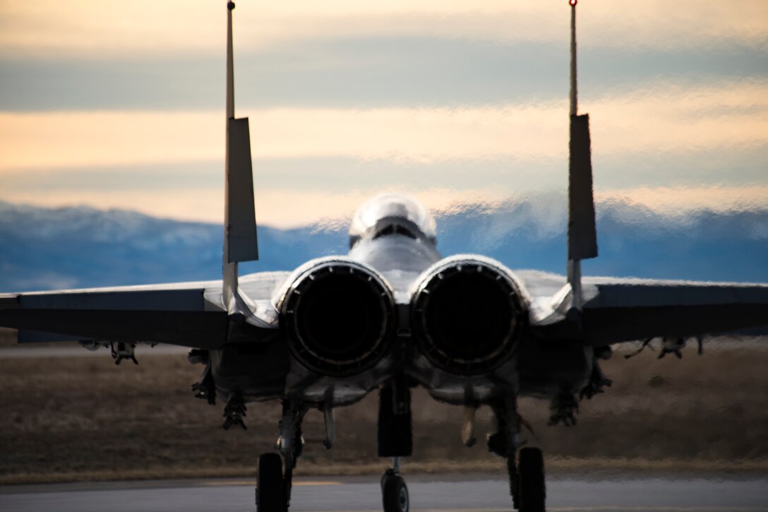 A military aircraft prepares to take off from a runway with mountains in the background.