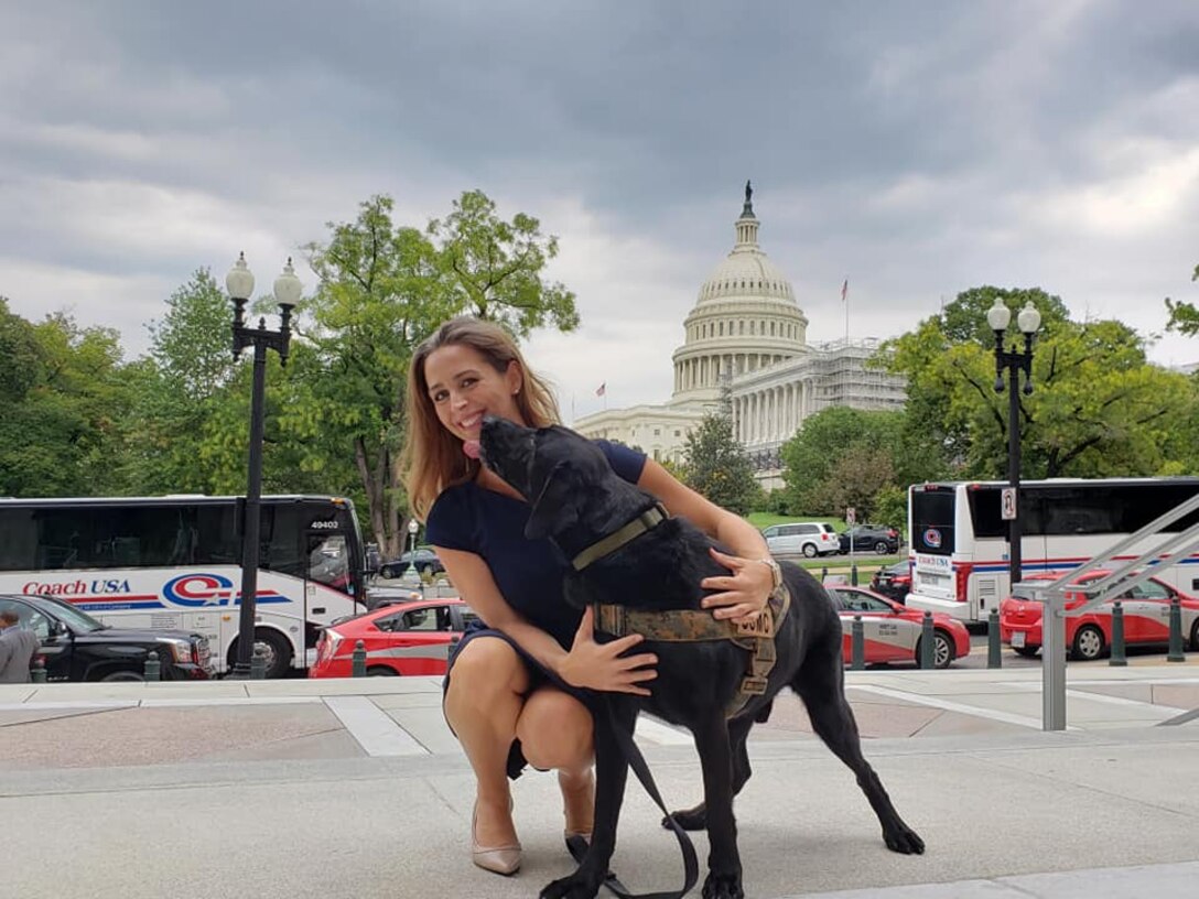 A black dog licks the face of a smiling woman as she squats down to hug the dog. In the background is the U.S. Capitol building.