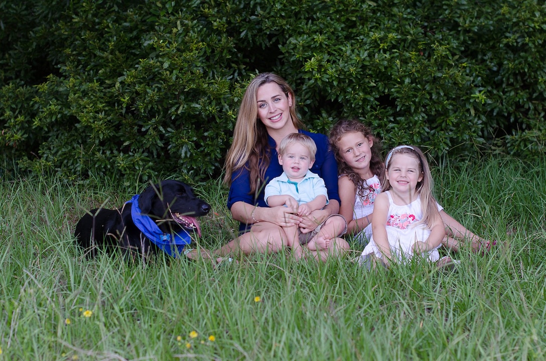 A woman and three children sit in grass beside a black dog wearing a blue bandana.