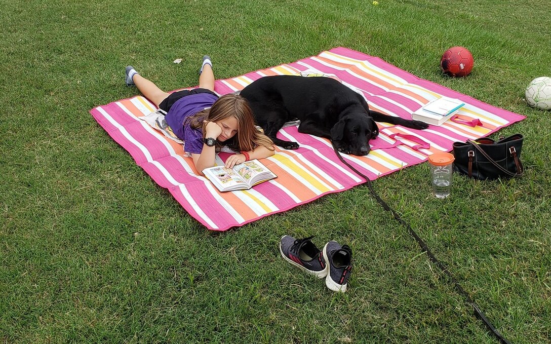 A black dog lays on a striped blanket with a young girl who is reading a book.