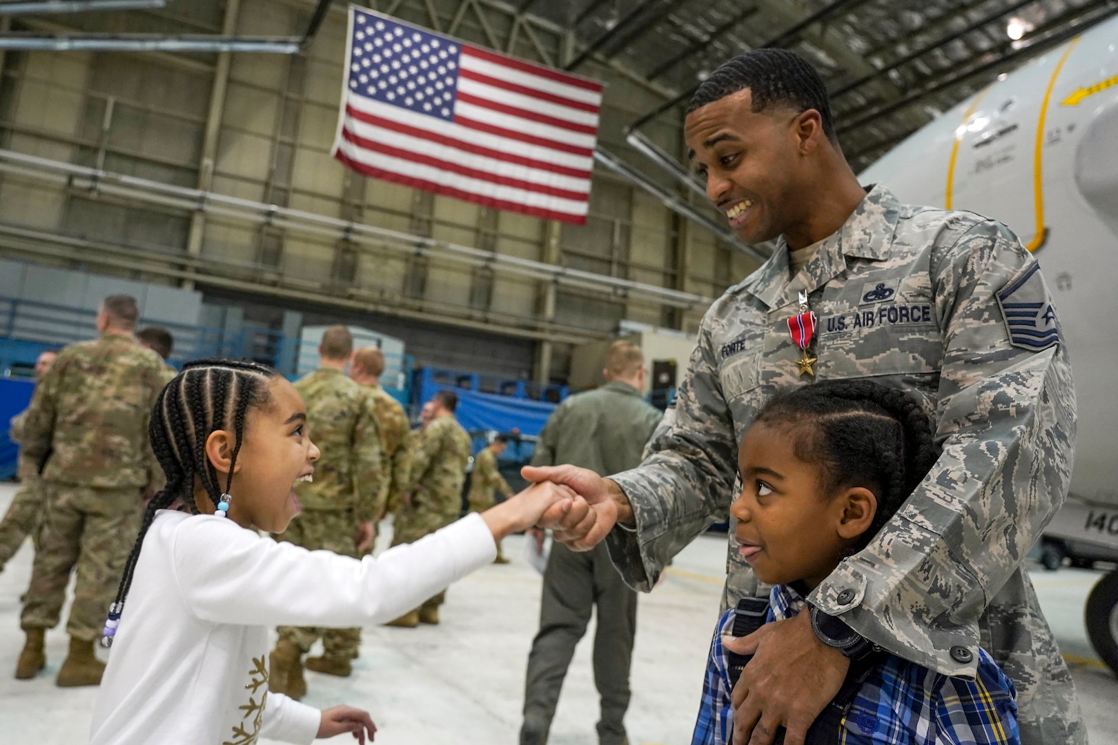 A smiling airman wearing a medal shakes hands with his smiling daughter while hugging his son in a hangar.