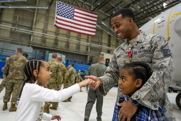 A smiling airman wearing a medal shakes hands with his smiling daughter while hugging his son in a hangar.
