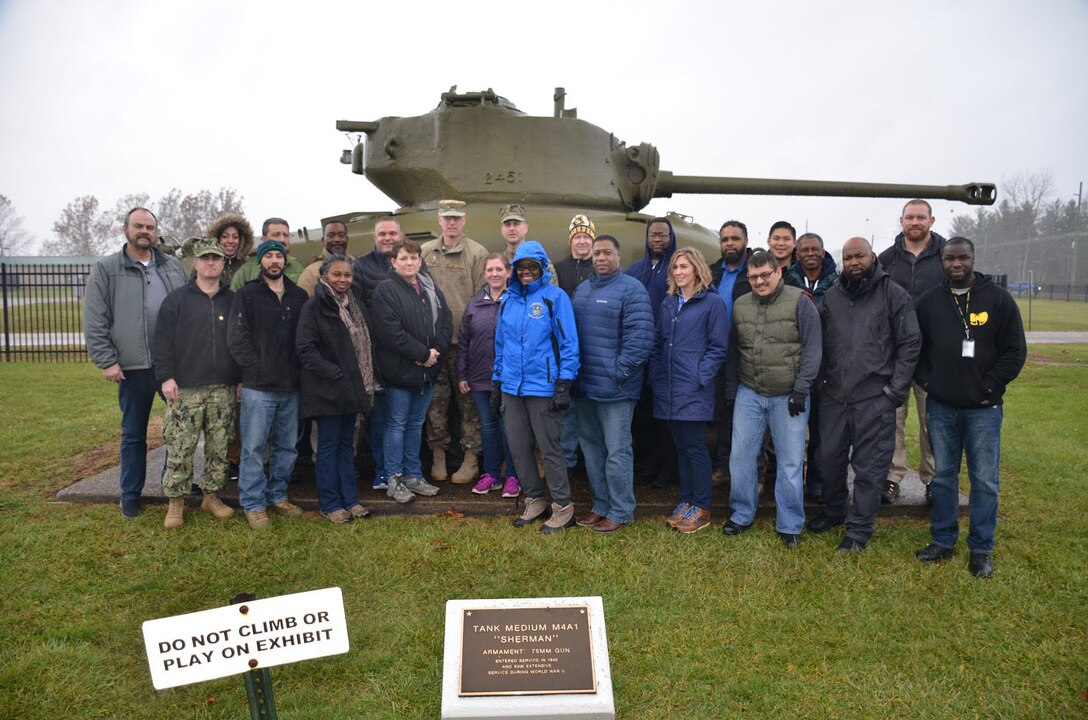 A group of men and women from the DLA RDT Red and White Teams pose in front of a tank during training at Camp Atterbury, Indiana.