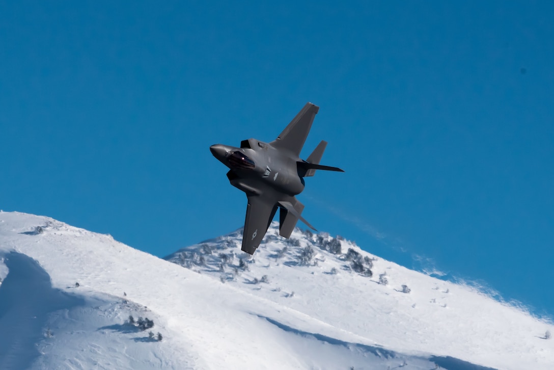 A military aircraft flies almost upside down above snowy mountains.