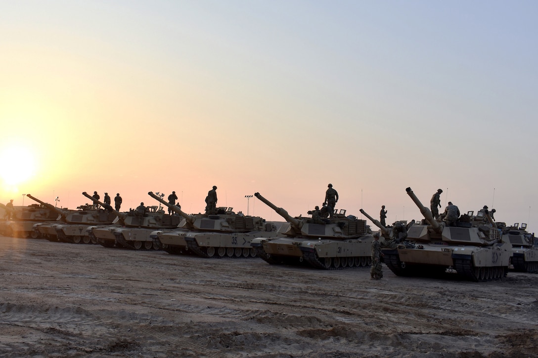 Armored vehicles sit in a row while soldiers climb on them at twilight.