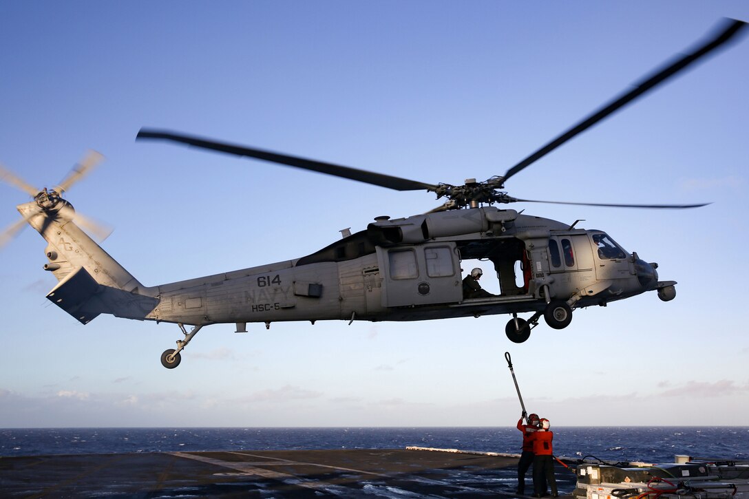 A military helicopter hovers above the deck of an aircraft carrier as sailors attach a load of cargo.