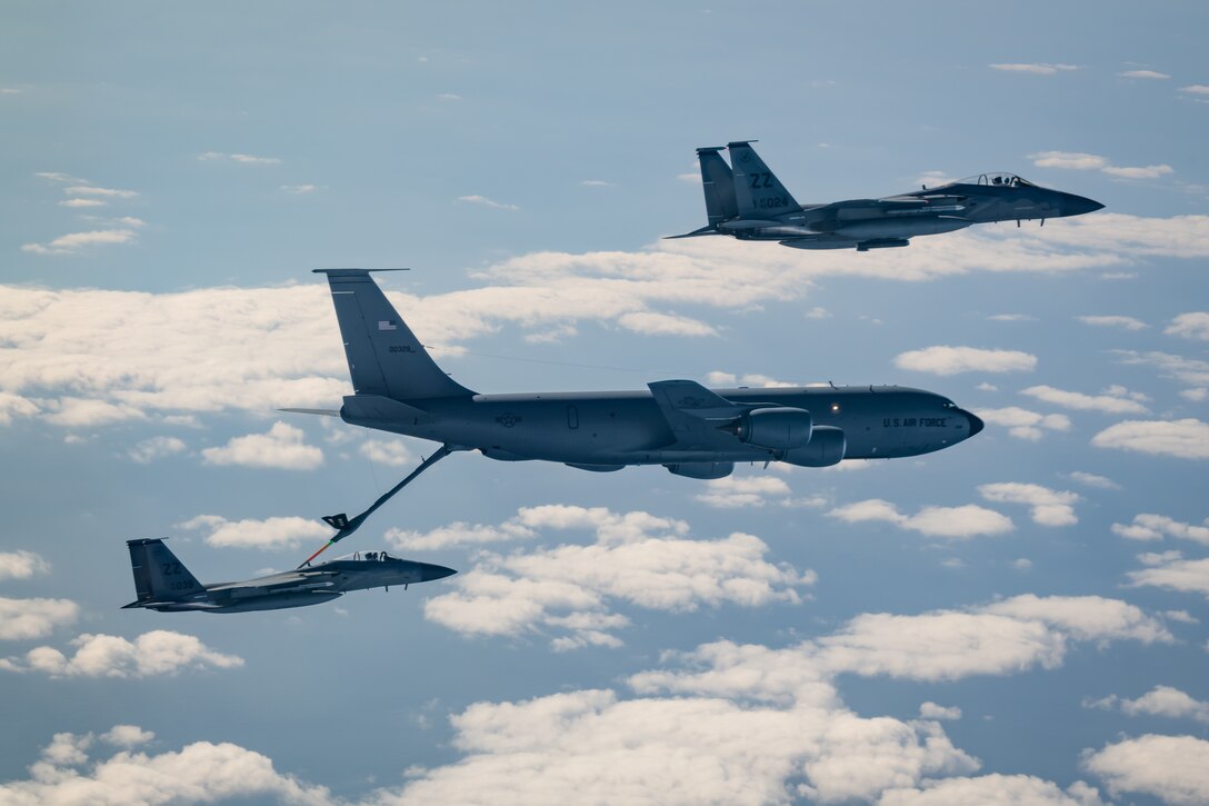 A large military aircraft fuels a smaller jet while a third jet flies above.