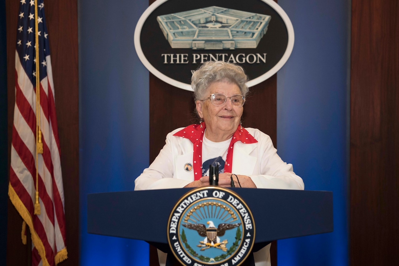 A woman stands at a lectern with the DOD logo.