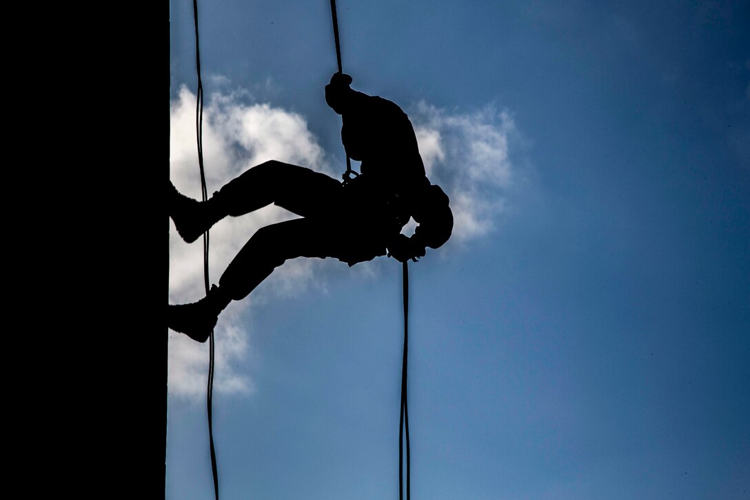 A Marine, shown in silhouette, rappels down a wall against a blue cloud-streaked sky.