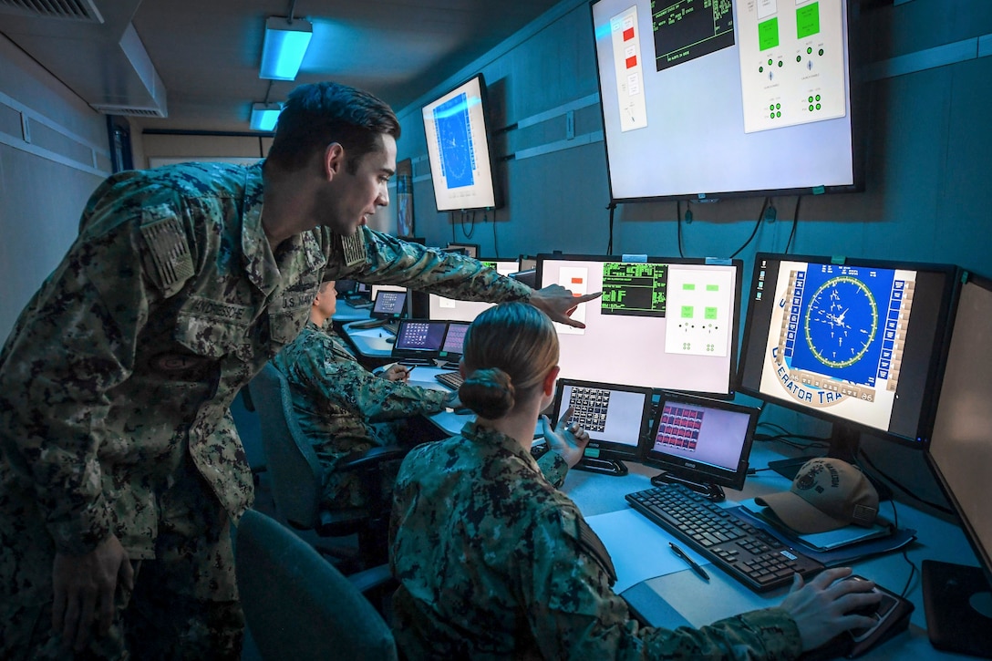 Sailors sit a consoles as an instructor points something out on a screen.