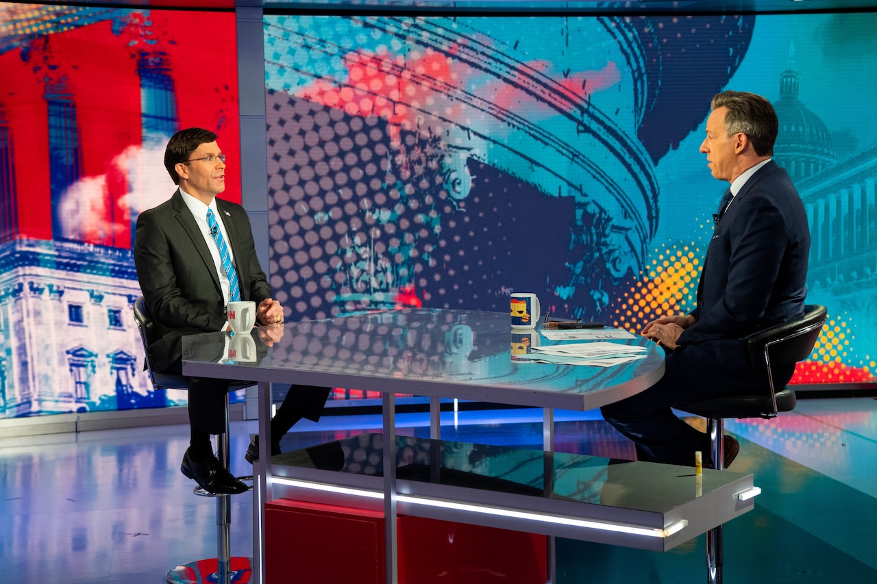 Defense Secretary Dr. Mark T. Esper sits across a table from another civilian inside a television studio.