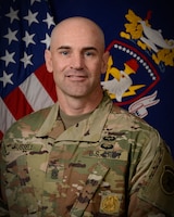 Photo of white man in green camouflage uniform standing in front of an American flag and the blue, yellow and white flag of the U.S. Army Recruiting and Retention College.