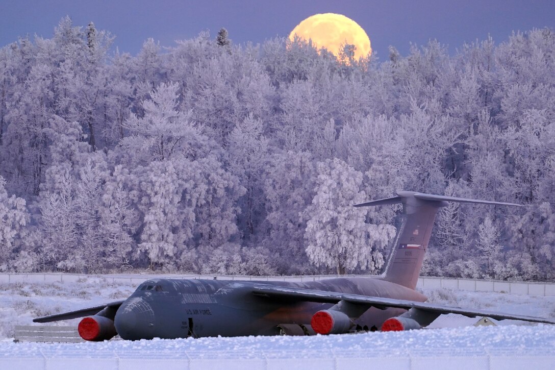 A large military aircraft sits on a runway surrounded by snow and  ice with the moon behind it.
