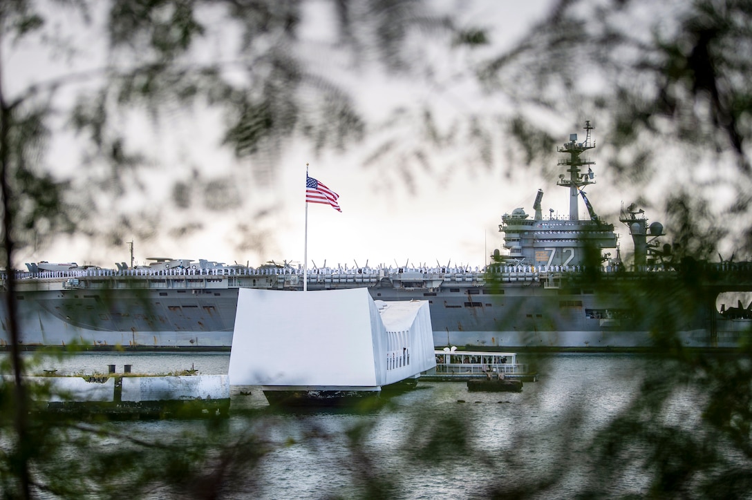 An aircraft carrier travels behind the USS Arizona Memorial, as seen through tree branches on nearby land.