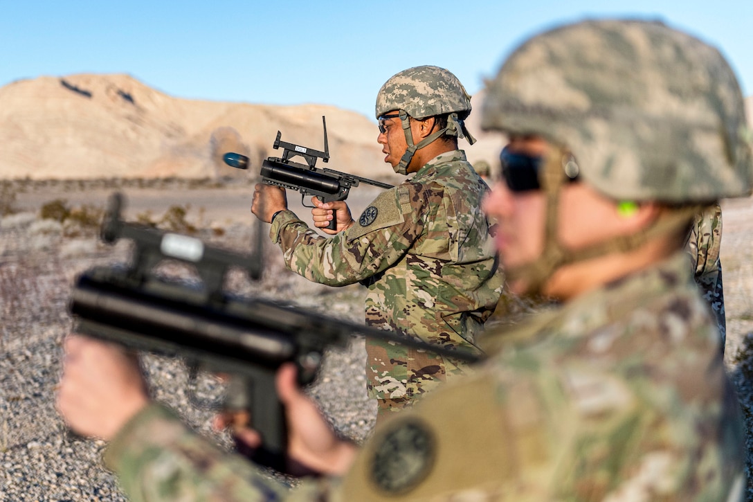 Soldiers stand outside in a row as one fires a training round from a grenade launcher