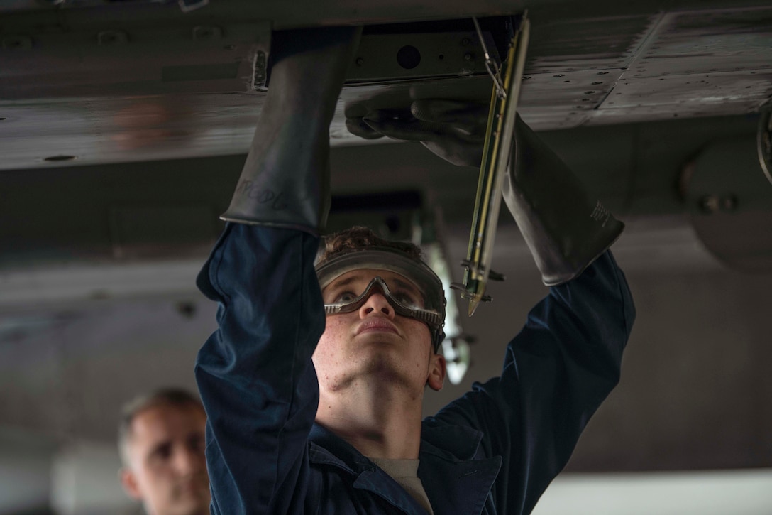 An airman wearing utility gloves and goggles looks up while working on an aircraft undercarriage.