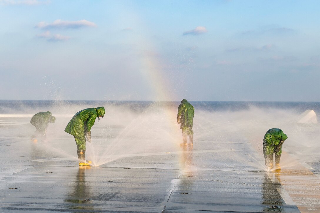 Sailors wearing green raincoats stand over sprinklers on a flight deck.