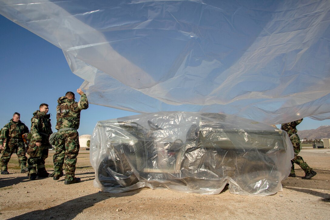 Airmen hold a sheet of transparent plastic over a vehicle already wrapped in a similar sheet.