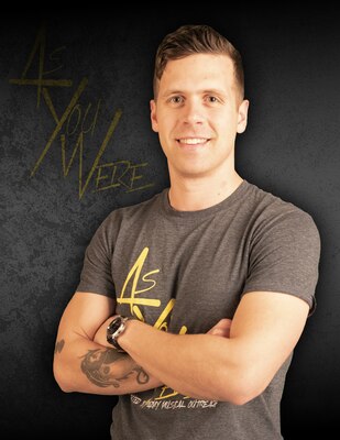 Brown-haired man with crossed arms and tattoo on right arm with gray t-shirt with yellow lettering against a gray background with yellow letters that say As You Were.
