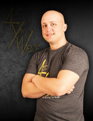 Bald, white man with crossed arms in grey shirt with yellow lettering against grey background with yellow words saying As You Were.