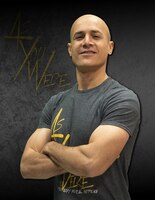 Bald male with arms crossed in grey t-shirt with yellow words with a black background and words that say As you Were.