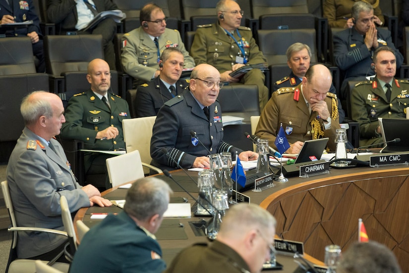Military officers from various nations sit behind microphones at a round table.