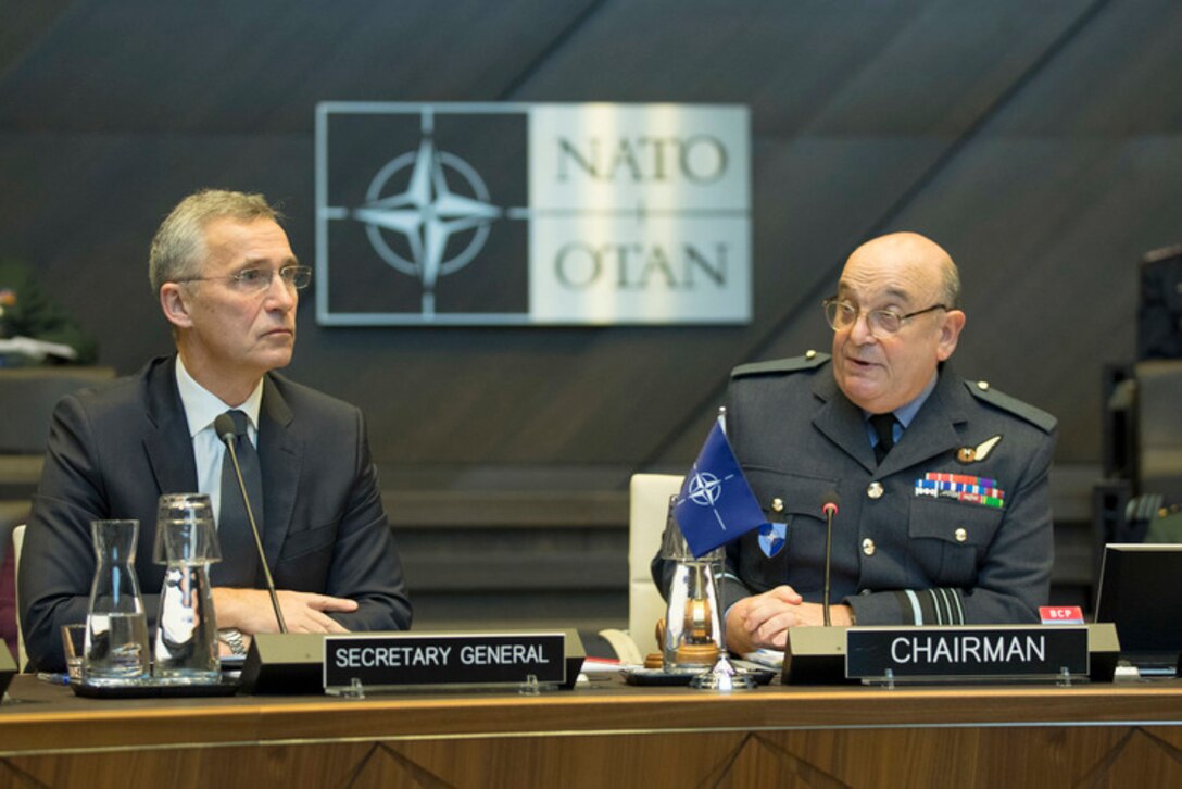 A civilian wearing a suit and a military officer in uniform sit behind microphones with a NATO emblem behind them.