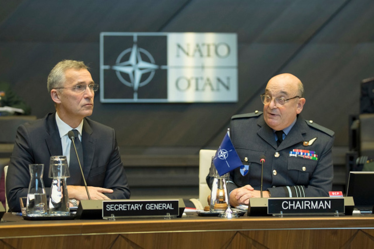 A civilian wearing a suit and a military officer in uniform sit behind microphones with a NATO emblem behind them.