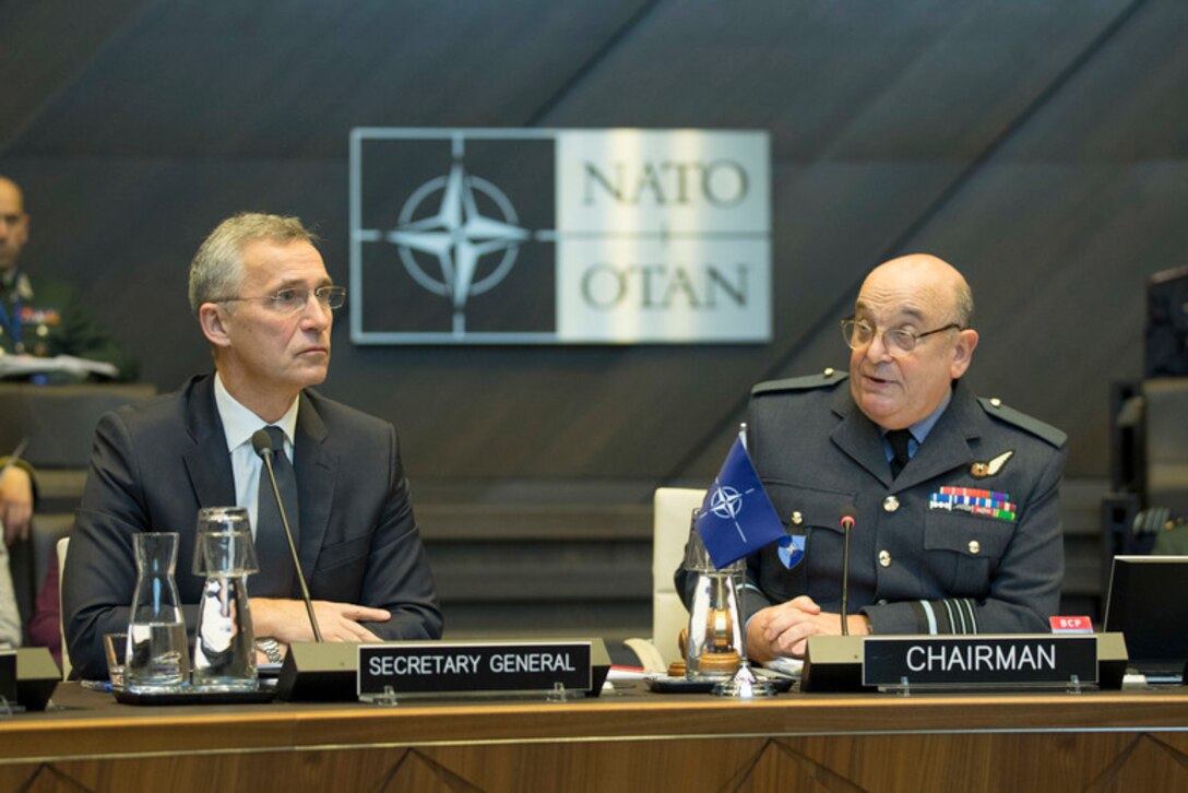 A civilian wearing a suit and a military officer in uniform sit behind microphones with a NATO emblem behind them.