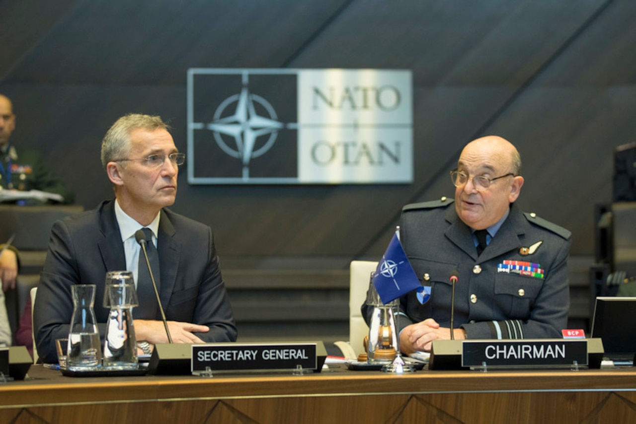 A civilian wearing a suit and a military officer in uniform sit behind microphones with a NATO emblem behind them.