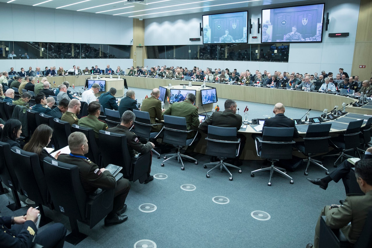 Wide-angle shot of a large conference facility, dominated by a round table with military officers seated around it.